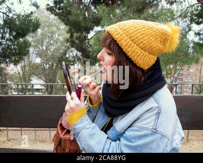 Young woman with yellow woolen cap sitting on a bench painting her lips in front of a mirror. Autumn urban concept. Stock Photo