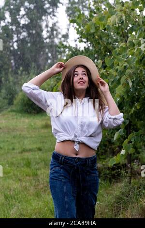 Summer warm rain. A young girl in a white shirt and blue jeans in the rain holds a hat on her head with her hands Stock Photo