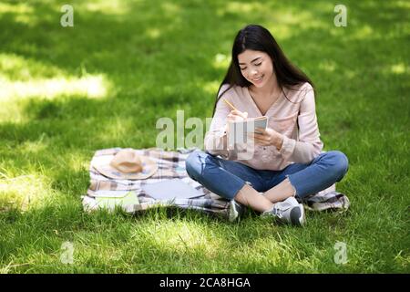 Beautiful asian girl sitting on plaid in park, taking notes to notepad Stock Photo