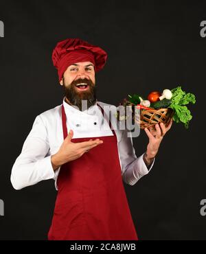 Man with beard on black background. Chef holds lettuce, tomato, pepper and mushrooms. Vegetarian diet concept. Cook with cheerful face in burgundy uniform presents vegetables in wicker bowl. Stock Photo