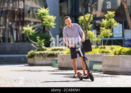 Young casual active sporty businessman in urban city center, wearing shirt and shorts, holding laptop bag, riding to work on electric scooter on a hot Stock Photo