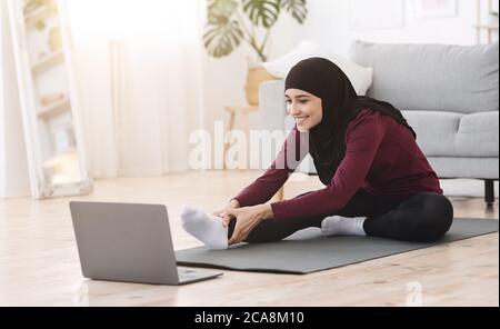 Home Pilates. Sporty arabic woman stretching leg muscles in front of laptop Stock Photo