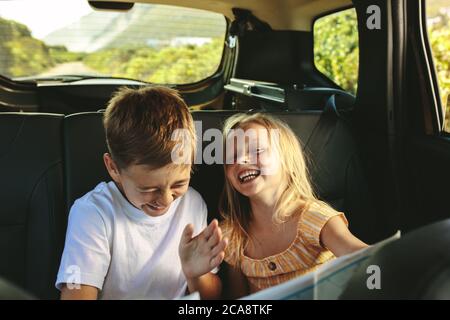 Siblings sitting on backseat of car looking at map and smiling. Kids traveling in a car on roadtrip playing with a map. Stock Photo