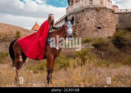 A beautiful girl with a sword in a medieval fantasy suit. A woman in a chain mail, cloak, crown, on a horse in combat ammunition. A girl riding a hors Stock Photo