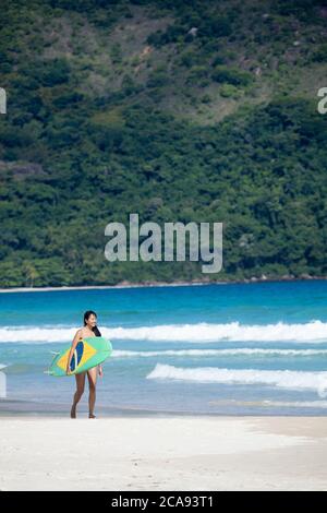 Beach shot of a Japanese Brazilian (Nipo-brasileiro) in a bikini carrying a surf board decorated with the Brazilian flag, Brazil, South America Stock Photo