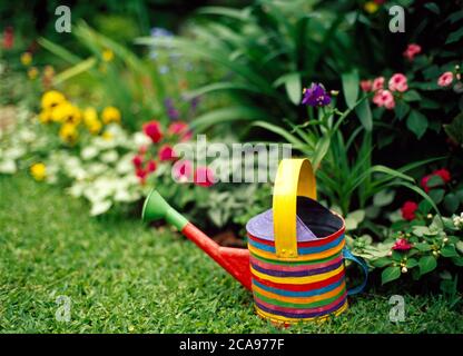 A handpainted striped watering can on lawn with a flowerbed Stock Photo
