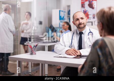 Young doctor looking worried at elderly aged patient during medical examination in hospital office. Young sick woman talking with mature medic on clinic corridor about treatment for her illness and nurse is holding x-ray. Stock Photo