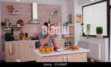 Freelancer drinking coffee and working on laptop during breakfast in kitchen. Entrepreneur in pajamas working on a laptop from home in kitchen. Stock Photo