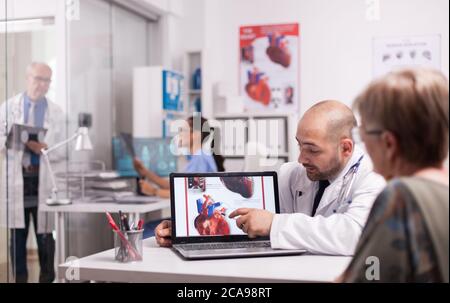 Young doctor pointing at heart on laptop screen in hospital office. Cardiologist with senior patient during consultation. Mature medic taking notes on clipboard on clinic corridor. Stock Photo