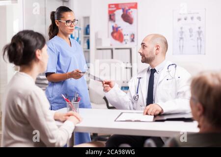 Nurse giving doctor patient x-ray during examination in hospital office. Daughter and her disabled mother in wheelchair at periodic medical check. Medic wearing white coat and stethoscope. Stock Photo