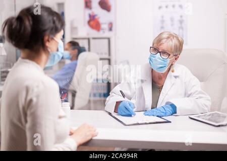 Doctor wearing protection mask against covid taking notes during consultation with patient in medical clinic. Nurse wearing blue uniform while working on computer. Stock Photo
