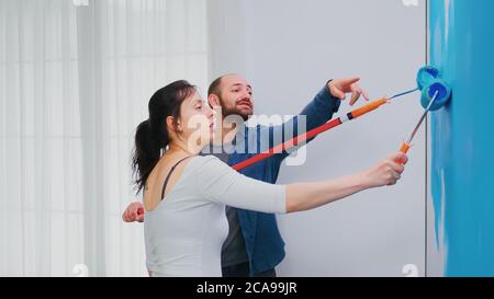 Couple repainting apartment wall with blue paint using roller brush. Apartment redecoration and home construction while renovating and improving. Repair and decorating. Stock Photo
