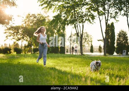 Stunning female caucasian dog owner enjoying summer walk with purebred pet in city park, having fun. Young happy woman wearing casual outfit laughing and running on grass with french bulldog on leash. Stock Photo