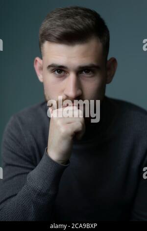 young bearded guy looks at the camera, on a gray background studio portrait. Psychological portrait. Handsome guy. Man of twenty five. Stock Photo