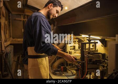 portrait of young caucasian handsome carpenter working, using powerful machine for wood cutting and sawing, man wear uniform. in wood furniture factor Stock Photo