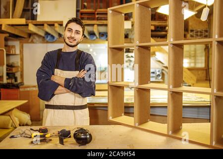 portrait of young caucasian positive carpenter at work place, handsome guy in work clothes smile and look at camera, enjoy being woodworker Stock Photo