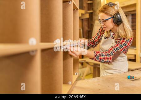 side view on attractive carpenter woman using tape-line during work, woman protect ears from noisy sounds in workshop Stock Photo
