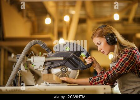 hardworking caucasian female carpenter manage wood cutting with the use of circular sawer, wearing headphones and uniform in workshop Stock Photo