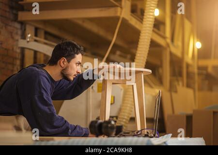portrait of young cerpenter checking handmade wooden chair after grinding in workshop. confident woodworker make furniture Stock Photo