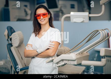 portrait of woman dentist in white doctor's uniform standing in office near equipment and look at camera, wearing red eyeglasses for safety. Medicine, Stock Photo