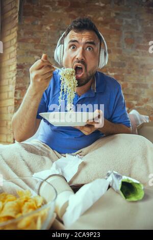 Shocked eating instant noodle, watching TV. Lazy man living in his bed surrounded with messy. No need to go out to be happy. Using gadgets, watching movie and series, looks emotional. Fast food. Stock Photo