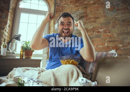 Crazy happy of favourite team. Videocall, social media. Lazy man living in his bed surrounded with messy. No need to go out to be happy. Using gadgets, watching movie and series, emotional. Fast food. Stock Photo