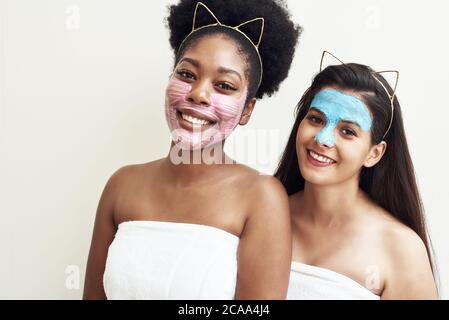 Multimasking. Two different ethnic young women are hugging. Women's faces are covered with pink and blue face masks. Facial beauty, facial care and spa concept. Stock Photo