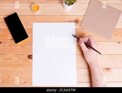 Hand holding the empty white paper sheet, and a calligraphy pen. Phone, plant envelop, and a small candle on a wooden desk. Stock Photo