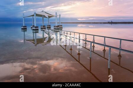 Calm morning at Ein Bokek Dead Sea beach, pink clouds reflection over water surface near steel rails leading to sun shade shelter Stock Photo
