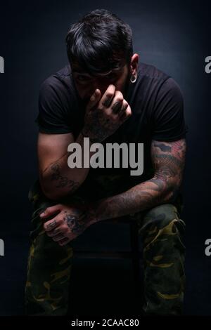 High contrast picture of a bearded male model sitting on a chair and covering his face with his tattooed hands Stock Photo