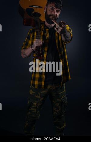 Tattooed man with a beard wearing a tartan yellow black shirt is  posing holding his guitar on his sholder and giving his middle finger Stock Photo