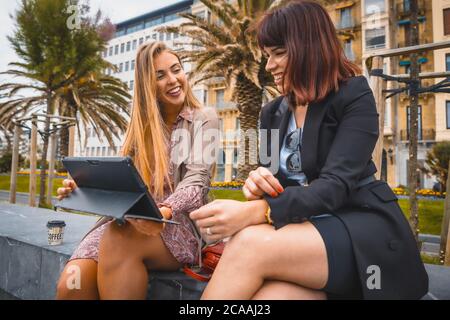 Horizontal shot of two young female entrepreneurs talking happily while looking at tablet Stock Photo
