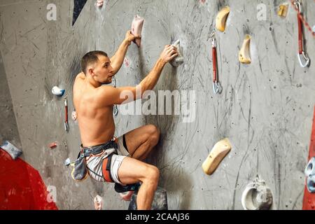 Unrecognizable athlete climber man exercising on grey practical wall with footholds, indoor shot, bouldering, rear view. Stock Photo