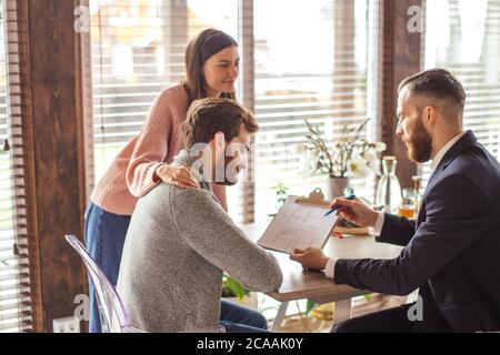 Family couple signing papers with realtor after concluding the realty purchase contract, sitting in the modern house Stock Photo