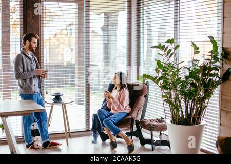 Young pretty couple in home clothes and slippers sitting at kitchen with large windows talking together about new project while drinking morning coffe Stock Photo