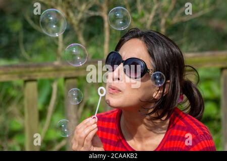Woman in red jumper, sunglasses and black long hair blowing bubbles outdoors Stock Photo