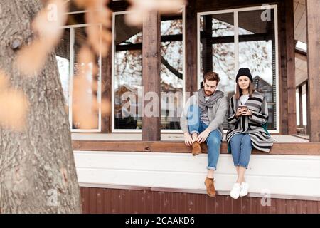 Young couple dressed in stylish warm coats sitting with cups on the terrace of the modern house enjoying beautiful fall view outdoors Stock Photo