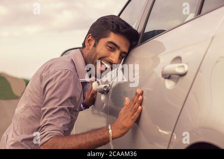 Young hispanic man wearing in formal shirt holding is petting his car and smiling outdoors. A new car, new driver concept Stock Photo