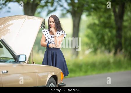 Woman in need for assistance desperately looking down to an open hood car engine after a breakdown. Stock Photo