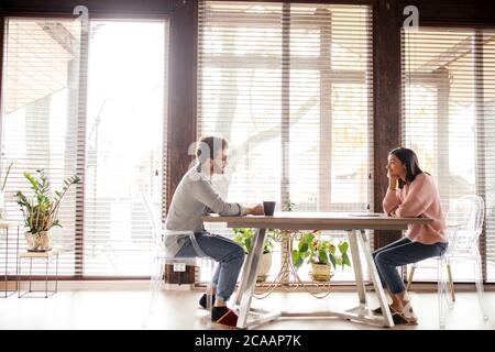 Panoramic view of big spacy room and caucasian couple sitting opposite at the same table in the house with big windows over background. Copy space. Stock Photo