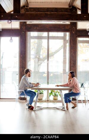Panoramic view of big spacy room and caucasian couple sitting opposite at the same table in the house with big windows over background. Copy space. Stock Photo
