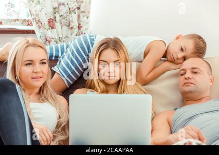 Adult parents with teen daughter and boy chilling on couch at home together and watching laptop Stock Photo