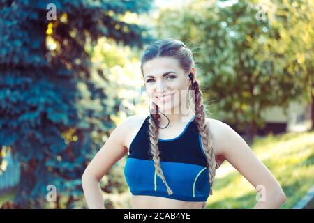 Young fit athletic woman in top and headphones standing in park and looking at camera Stock Photo