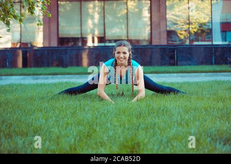 Pretty young brunette in sportswear stretching legs on green meadow standing in splits and smiling at camera Stock Photo