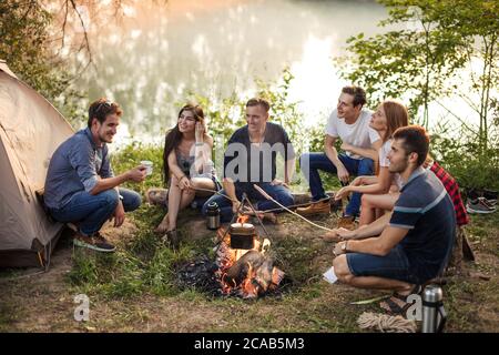 young attractive people holding sticks with sausages on the bonfire. process of preparing meal in nature. young people are huddling around the campfir Stock Photo