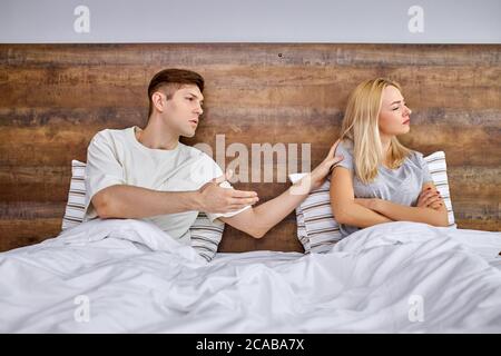 young caucasian man try to calm his girlfriend, resolve situation and talk with her, she lie on bed turning away from him, offended woman Stock Photo