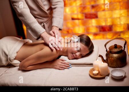 Professional and successful masseur doing massage on shoulders of young lady lying down on desk in spa salon Stock Photo