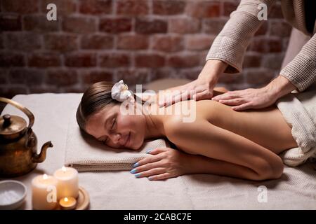 Beautiful young lady lying down on her belly in spa salon, getting massage on her back, relaxes woman with flower on head, candles near her. Stock Photo