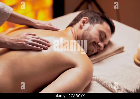 Masseur doing back massage to young caucasian man lying on spa table and relax with closed eyes, yellow spa room background Stock Photo