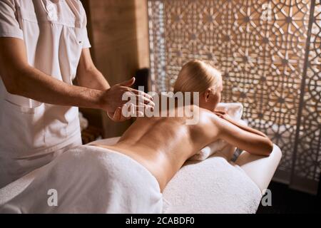 Brutal hands of male professional masseur rubbing oil, making preparation for giving hand massage to young blonde girl lying on massage table in welln Stock Photo